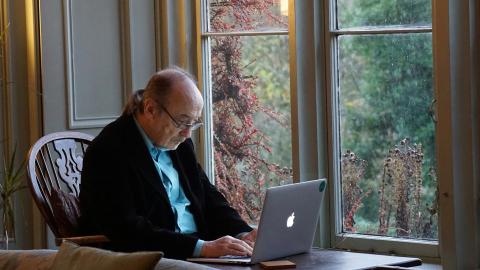 An older person in glasses looks at a laptop while sitting inside a house near a window.