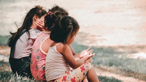 Three young girls sit on a grassy hillside in a smoggy city.