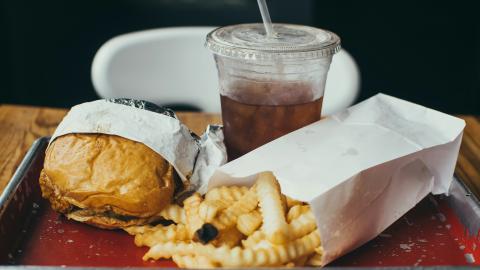 Photo of a tray on a table with a hamburger and fries in packaging, and a plastic cup of soda with ice and a straw.