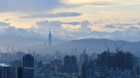 The skyline of Taipei showing skyscrapers, with a mountain and hazy sky with clouds in the background.