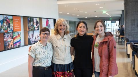 Four women (Orly Stampfer, Catherine Karr, Esther Min and Hanna Bailey) stand together in front of art about public health.