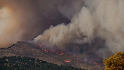 A smoke plume rises from a wildfire on a hillside near a group of houses.