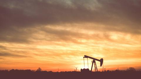 An oil drilling rig backlit by smoky skies at sunset with windmills in the background.