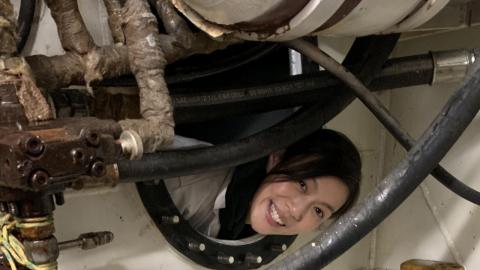 Photo of Amy Liu poking her head through an opening inside a ship, surrounded by plumbing pipes and tubes.