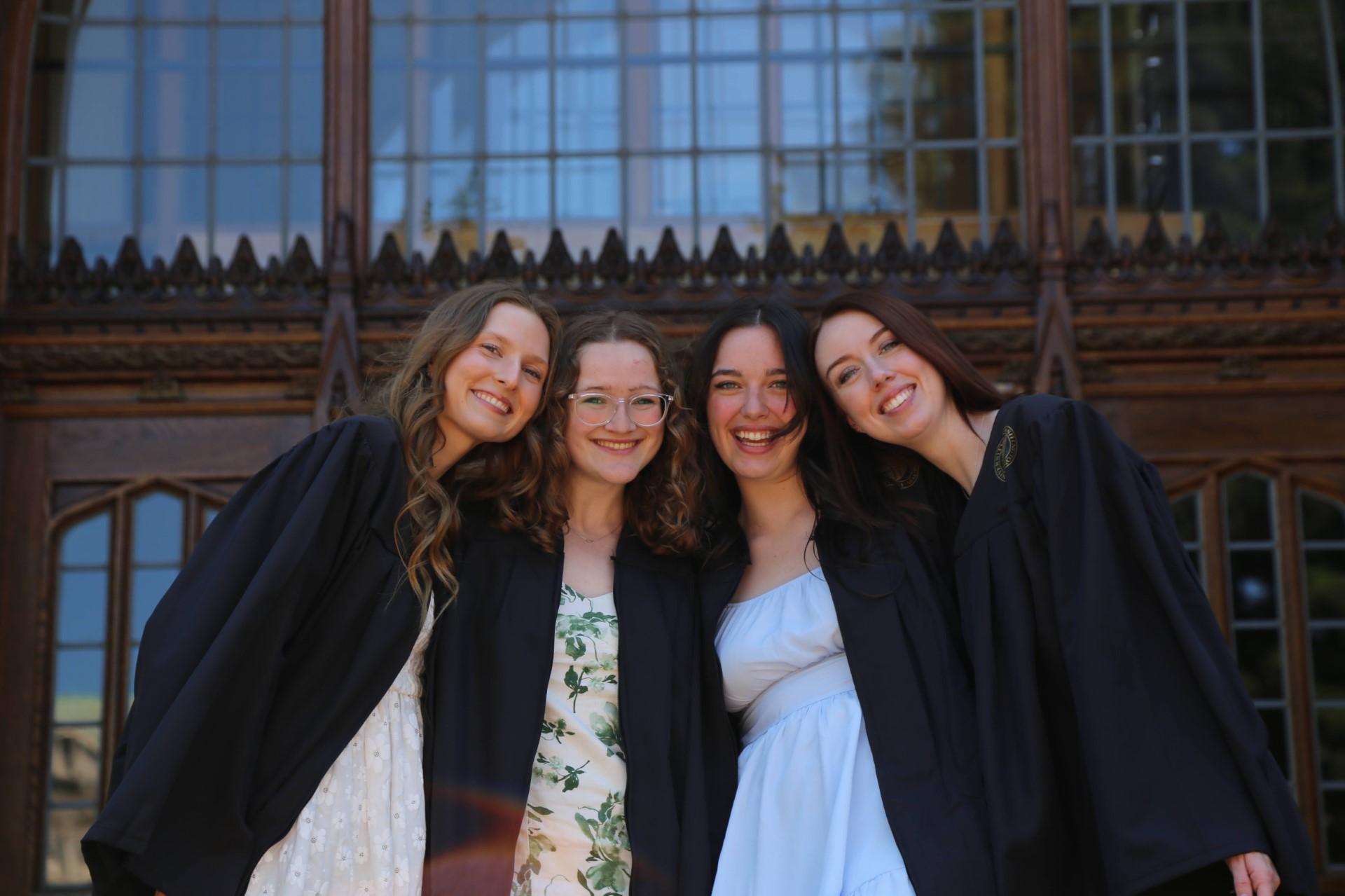 Four young women pose in black graduation robes in front of a library.