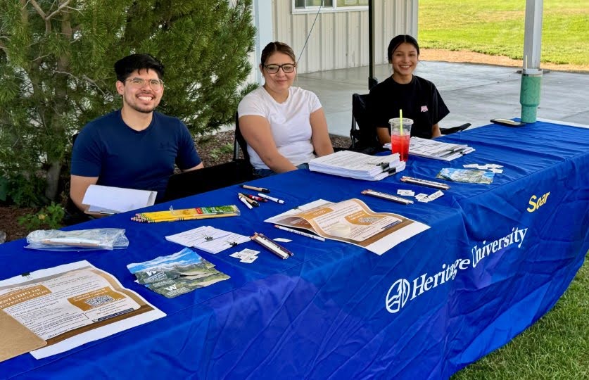 Three people sit behind a table outside covered with a banner from Heritage University, with materials for sharing with people at a information fair.