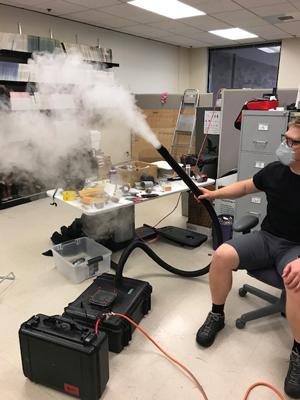 A seated researcher holds a fogging machine with a black tube emitting fog.