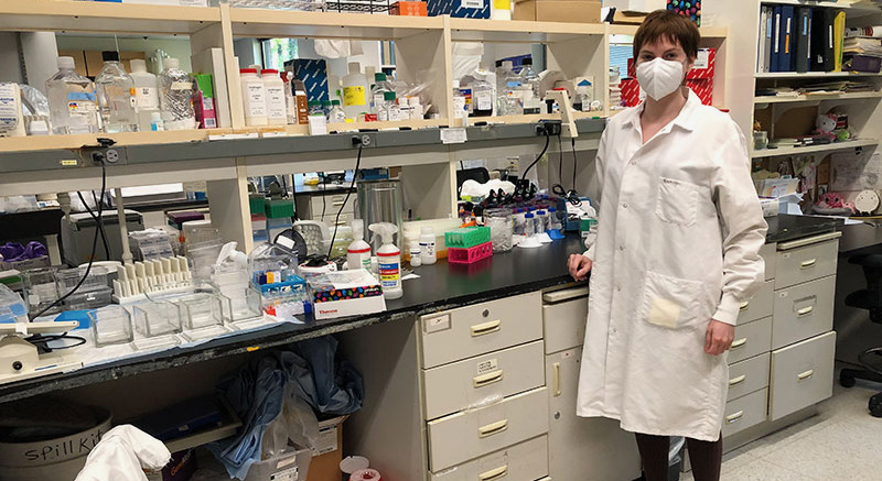 Jackie Garrick stands in front of a cluttered lab bench, wearing a white face mask and lab coat. 