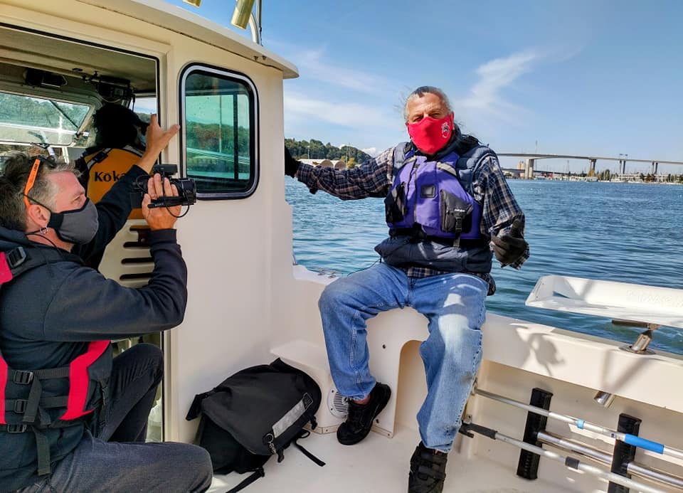 James Rasmussen in face mask and life vest sitting on boat while speaking about the Duwamish River with a film crew off to the side