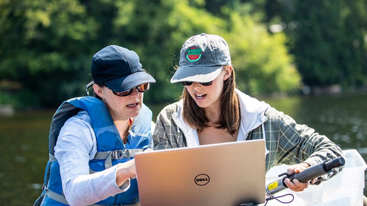 Dr. Neumann points at a laptop with graduate student, Samantha Fung, on a boat in a lake field site.  
