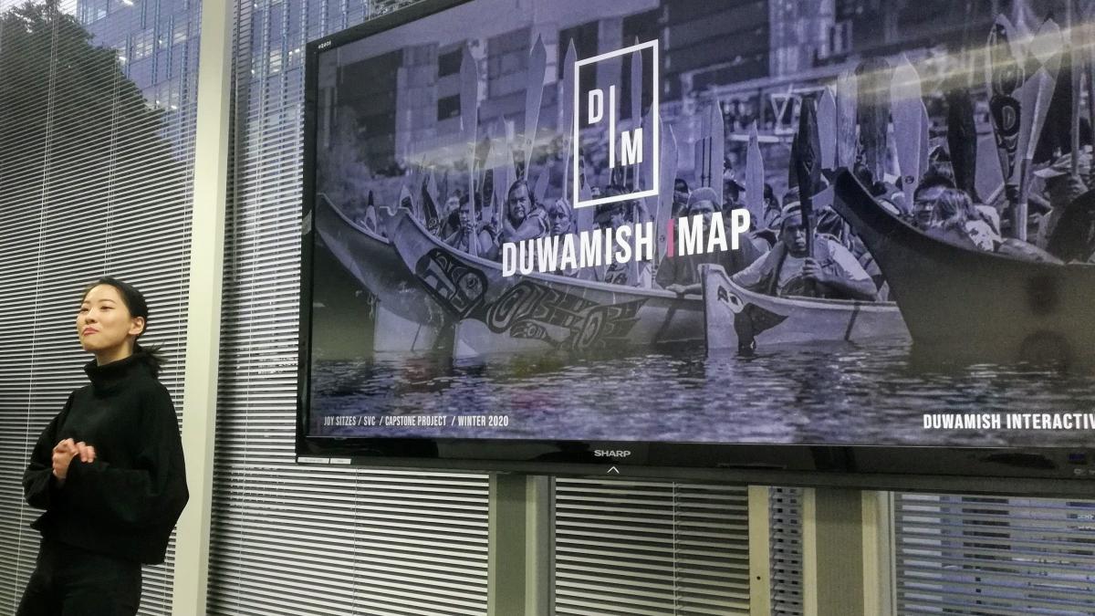 A young woman in black poses in front of a screen displaying a title slide for a virtual map of the Duwamish River.