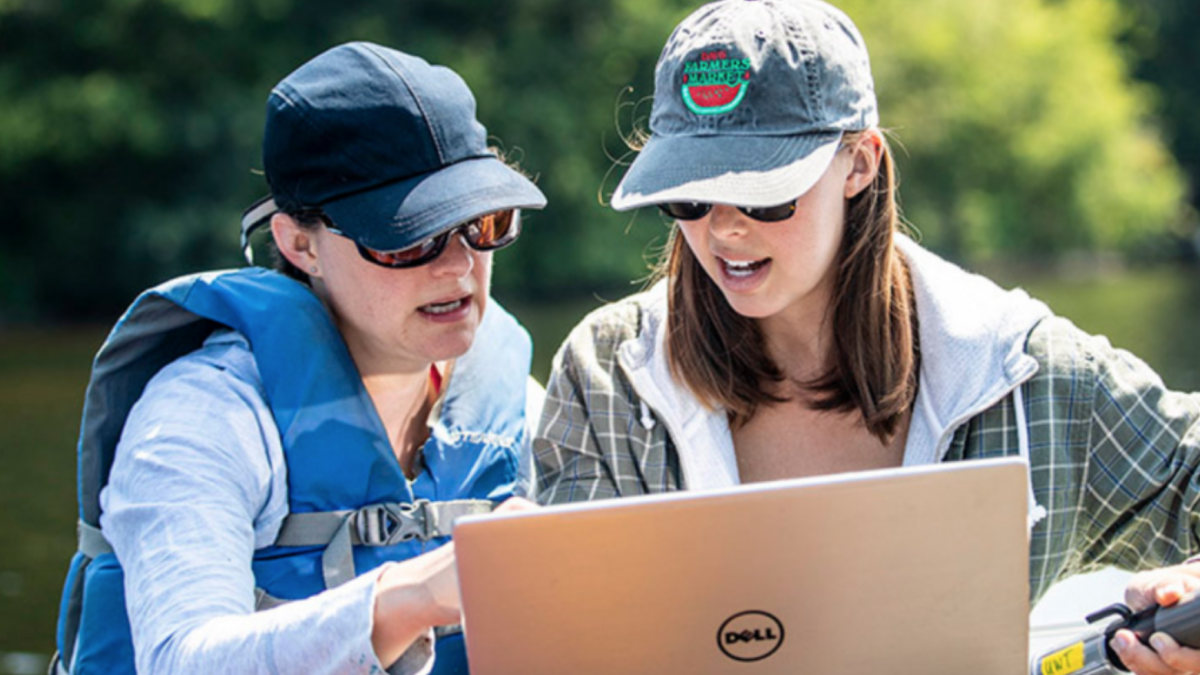 Two researchers look at a laptop together by a lake