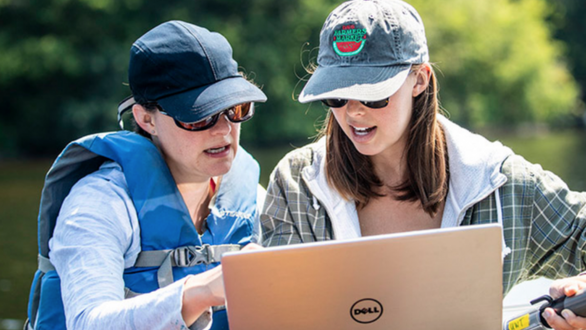 Rebecca Neumann and Samantha Fung look at a laptop together in a boat on a lake