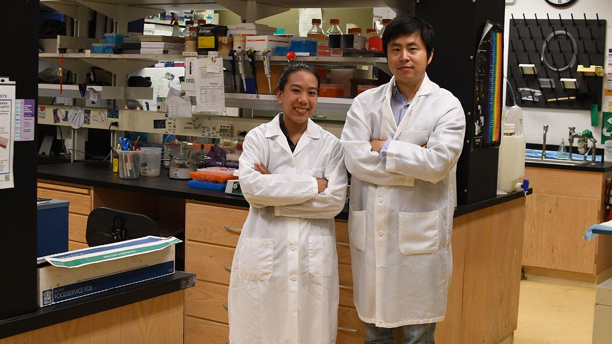 Dr. Wang (right) poses with fellow UW SRP trainee Megumi Matsushita in the Xia lab. Both researchers wear white lab coats.   