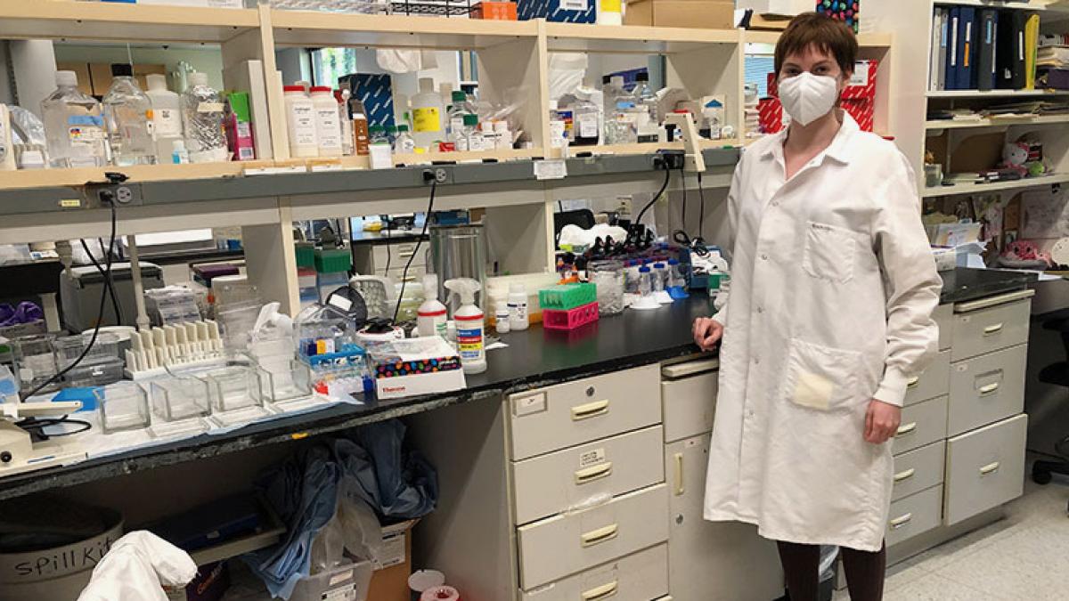 Jackie Garrick poses in a white lab coat in front of a lab bench with equipment and shelves of bottles